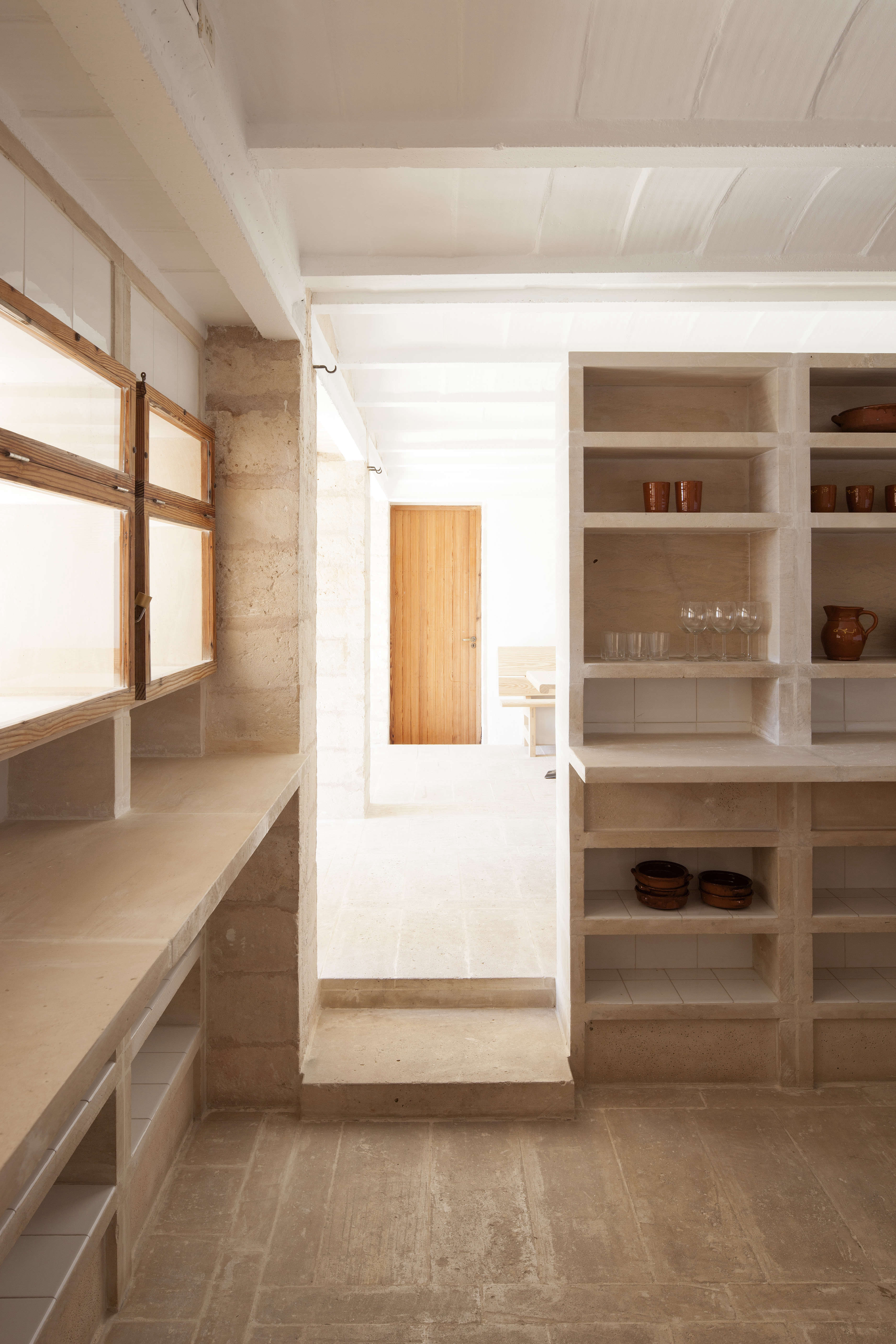 A concrete kitchen of open shelving, tiled under-the-counter shelves, and spare kitchenware.